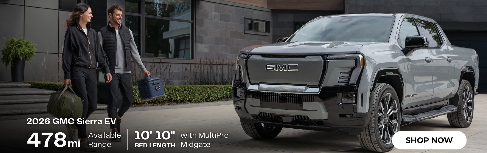 A couple with luggage walks towards a gray 2026 GMC Sierra EV pickup truck parked outside a modern building.