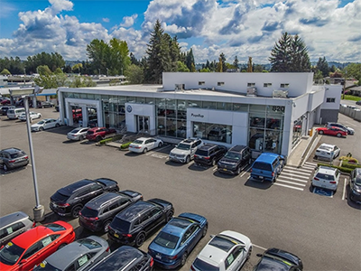 Aerial view of a white car dealership building with a large parking lot full of cars under a partly cloudy sky.
