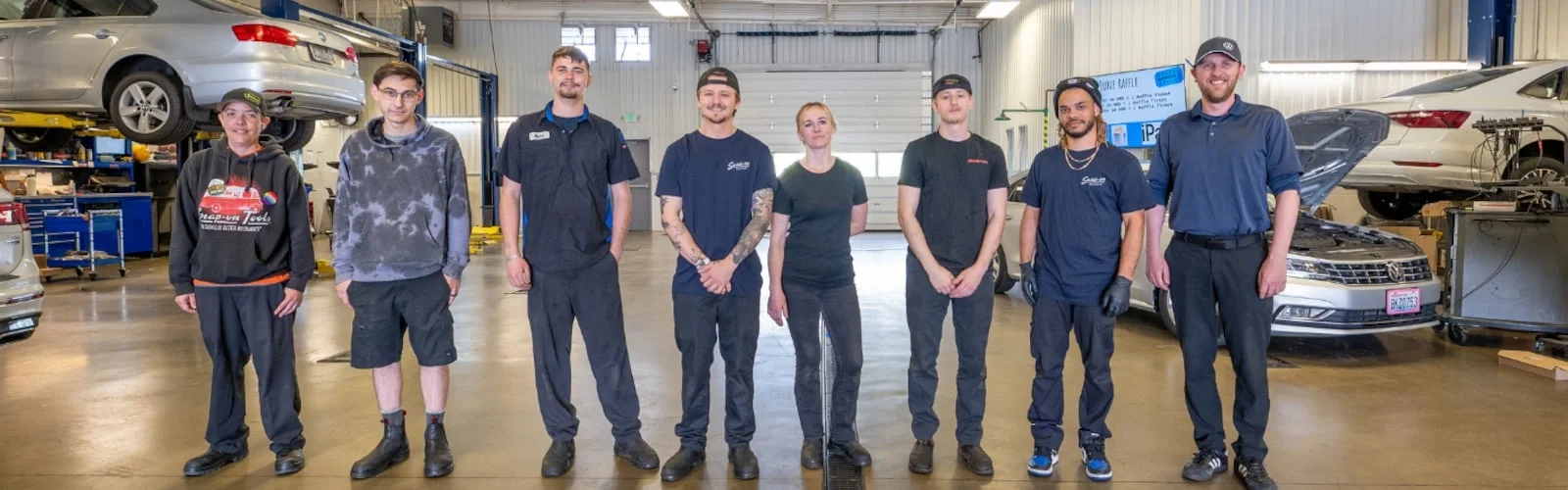 Eight smiling auto technicians, seven men and one woman, stand in a brightly lit garage with cars on lifts.