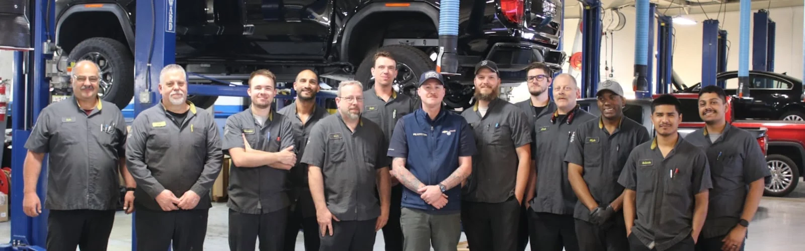 A team of 14 male auto mechanics in uniform stand smiling in a well-lit garage with vehicles on lifts.