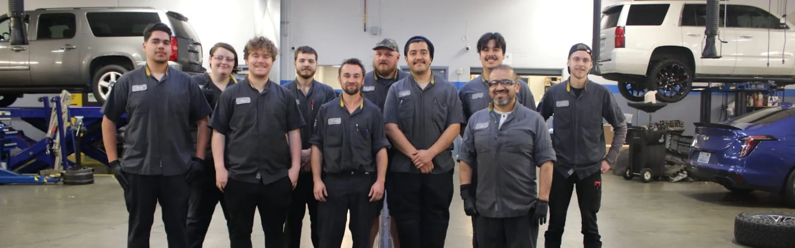 A diverse group of ten mechanics in gray uniforms smiles while standing in a busy auto repair shop.