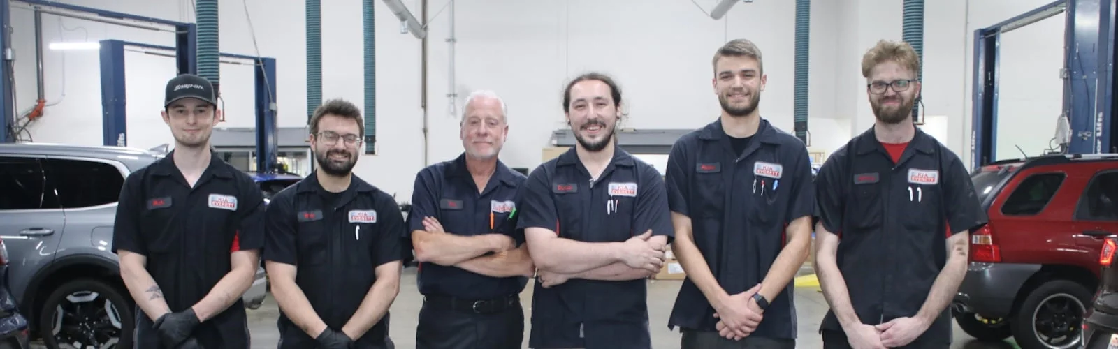 Six smiling mechanics, one grey-haired, pose in black uniforms with cars in the background of a garage.